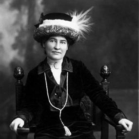 Black and white, three-quarter-length photo of Willa Cather, c. 1912, sitting facing the camera with her hands on the arms of her Renaissance-style  chair. She is wearing a hat with a turned-up brim holding a plume of white feathers; her blouse and jacket are black. She has a short pendant at her throat, and a long, tasselled necklace which came to her from Sarah Orne Jewett.
