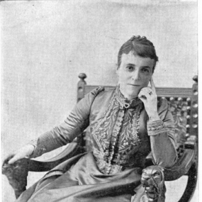 Black and white photograph of Laura Ormiston Chant, seated in an ornately carved rocking chair with one elbow resting on an armrest,  her arm upright and her head propped against that hand. Her other arm is outstretched so that hand can rest on the other armrest. She wears a dress with a pattern of stitched vines and buttons up the front, and her hair is pulled back.