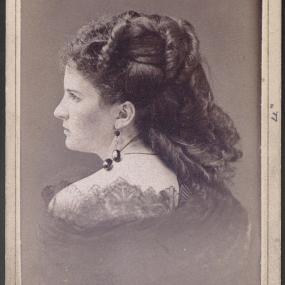 Sepia head-and-shoulders photo of Kate Chopin, taken from behind but showing her face in profile. Her hair is in ringlets, some pinned to her head, some at her back. She is wearing a necklace and earrings of dark stones, and a dress with lace collar.