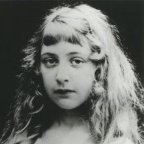 Black and white photo of Agatha Christie as a child. Her hair is in curls and her face is amusingly expressionless. It is unclear what she is wearing, however we believe it is a dress, because of the fabric visible on her shoulder.