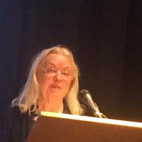 Somewhat blurry colour photo of Gillian Clarke standing at a lectern with a microphone. She is wearing something dark, with glasses, and has loose shoulder-length blonde hair.
