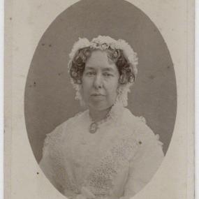 Portrait of Mary Cowden Clarke, circa 1870's. Black and white portrait of Mary Cowden Clarke. She faces forward with a neutral expression, and her hair is covered by a lacy bonnet, save for a few curls at that fall just above her cheekbones. She wears a white dress with lace detailing.