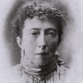Portrait of Agnes Mary Clerke. Black and white portrait of Agnes Mary Clerke. She faces forward with a neutral expression and her hair is short, pulled away from her face. Though the photo appears to be slightly damaged along the bottom, her face and neck are framed and visible. She wears a shirt with a high collar, and what looks like a pendant necklace, though the damage to the photo makes what is on the pendant unclear.