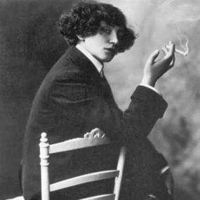 Black and white photograph of Colette wearing a man's suit (with cuffed jacket) and dress shoes. She is sittine turned in a chair so that her arm rests on its back, crossing her left leg over her right, and smoking a cigarette. She wear her hair in a curly bob.