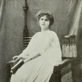 Black and white photo of Mary Corelli by F. Adrian, published in 1904. She sits in a chair that is adorned in unusual style with tassels, wearing a pale dress and holding an open book. Her hair is short and curly, artfully disarranged.