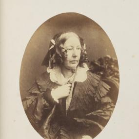 Sepia-toned albumen print of Louisa Stuart Costello by Herbert Watkins, 1857. She sits on a carved chair, gazing thoughtfully to her left, with her right hand up to her neckline. Her hair is in ringlets in front and decorated with bows. She wears a dark dress with ruffles descending the sleeves, and a white lace collar. National Portrait Gallery.