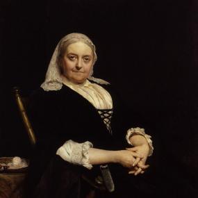 Oil portrait of Dinah Mulock Craik, by Sir Hubert von Herkomer, 1887. She sits in a wooden chair, her full-figured torso facing to her left but her face turned to look directly at the viewer and hands folded in her lap. She wears a dark dress which merges with the dark background, and a white high-collared blouse with embroidery at the sleeves and lacing in front. She wears a lace headpiece hanging from her grey hair, and her expression is stern. National Portrait Gallery.