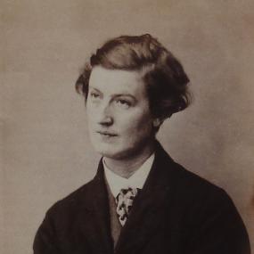 Sepia toned photograph of Hannah Cullwick, shown from the waist up. She is slightly turned, with her arms crossed, and gaze into the distance with a slightly challenging expression. She is wearing an oversize suit jacket, a collared shirt and cravat; her hair is cut short.