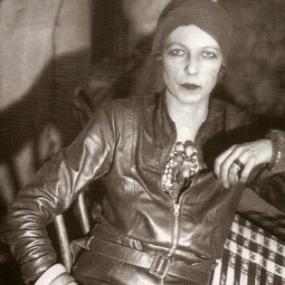 Black and white photograph of Nancy Cunard. She is seated in a casual pose with elbows out, facing the camera, wearing a belted leather jacket, what looks like a cloche hat, and her trademark stack of African ivory bangles.