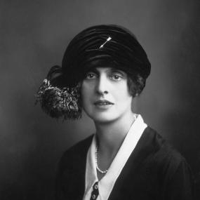 Black-and-white portrait of E.M. Delafield, wearing a cloche hat that gathers at one side with what looks like animal fur and an arrow brooch
            in the centre, 1922.