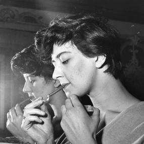 Black-and-white portrait photo of Shelagh Delany in profile, standing in front of a mirror and lighting a cigar, 1950. 