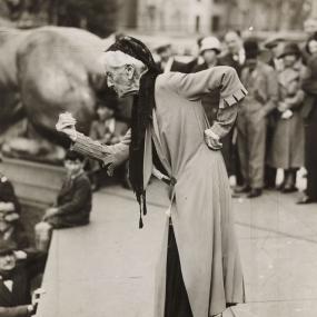 Black-and-white photo of Charlotte Despard speaking at an anti-fascist rally in at Trafalgar Square in London, c 1930s. She is kneeling
            forward with her left arm and her waist and her right arm hooked forward in a fist.