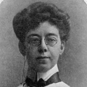 Black and white, head-and-shoulder photograph of Mary Angela Dickens looking straight at the camera. Her dark hair is short and curly; she has glasses perched on her nose, and her dress is masculine: a white shirt with a high, stiff collar and a dark bow tie outside it.