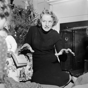 Black-and-white photo of Monica Dickens, circa 1954, reading her grandfather's "A Christmas Carol" to her children. Her hands are both
            reaching out and behind her is a Christmas tree and an unlit fireplace.