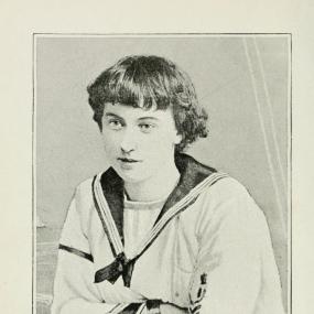 Black and white photo of Florence Dixie, used as frontispiece to her "Gloriana", 1890. She leans on a wooden railing, facing the viewer, wearing a sailor suit (white blouse with collar like a neckerchief. Her hair is short, with a short fringe. Beneath, her signature is reproduced: "faithfully yours, Florence Dixie."