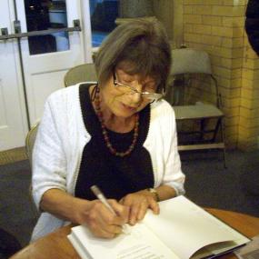 Photo of Margaret Drabble signing a book at Beverley Bookfest in Yorkshire on 15 October 2011. She wears a white cardigan over black, a necklace, and glasses.