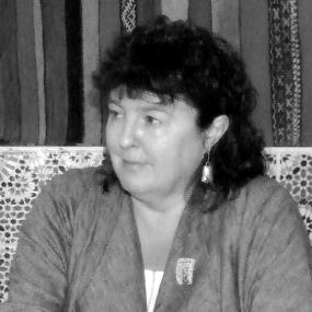 Black and white photograph of Carol Ann Duffy at Humber Mouth literary festival, 2009. She sits with hands folded on a table that is not pictured. She wears a loose jacket with a large lapel, a brooch, and earrings. She looks to her right with a neutral expression, and her hair is curly, falling loose around her shoulders and forehead.