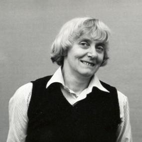 Black and white, half-length photograph of Maureen Duffy, her head slightly on one side and smiling. She wears a dark pullover with a striped shirt; her grey hair is in a page-boy cut with a fringe.