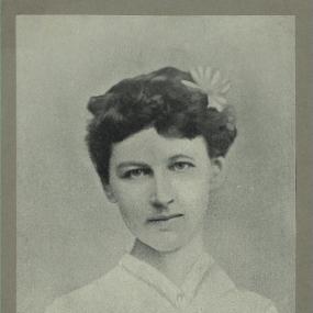 Black and white, head-and-shoulders photograph of Sara Jeannette Duncan, wearing a light buttoned blouse with a white flower in her dark curly hair.