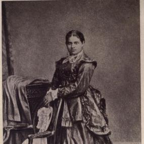 Black and white photograph of Toru Dutt, 1921. She stands holding a chair back in front of what looks like a wooden dresser, turned to her right but looking straight at the camera. She wears a heavy ruffled gown; her hair is parted in the middle and pulled back.