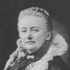 Black and white portrait photograph of Amelia B. Edwards, signed "Yours very sincerely, Amelia B. Edwards", 1890. She sits facing the viewer, turned slightly to her right. She wears a coat lined with fur, and a scarf over a textured blouse. Her hair is pulled away from her face, and she smiles slightly.