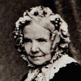 Black and white photograph of Charlotte Elliott. She sits at a slight angle to the camera, looking right. She wears a dark dress with white lace detail at the collar, and a white cap which covers most of her hair, with a few tight curls framing her face.