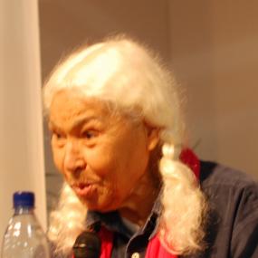 Colour photo of Nawal El Saadawi at the Göteborg Book Fair in Sweden, 2010. She is leaning forward speaking animatedly into a microphone, wearing a blue jacket and a thin pink scarf. Her white hair is tied in two long  bunches.