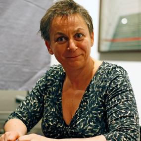 Photo of Anne Enright sitting at what appears to be a signing with a marker in her right hand. Her hair is cut short and is auburn, and she is wearing a green and dark green floral patterned top. In the background, there is a mirror on the wall.