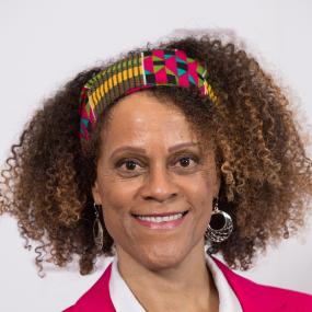 Close-up colour photograph of Bernardine Evaristo, during the Booker Prize Winner event at Guildhall, London, 14 October 2019. She wears a
            colorful patterned headband, silver hoop earings, and a bright pink blazer jacket.