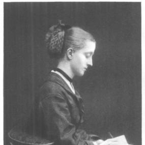 Photo of Julia Horatia Ewing in profile, sitting in a chair writing in a small book. Her hair is pulled tightly back and plaited in an ornate bun. Her dress is dark, with a glimpse of white at throat and wrists.