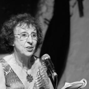 Black and white photograph of Elaine Feinstein by Kaido Vainomaa, reading at Shaar International Poetry Festival in Tel_Aviv, 2010. She is seen from the waist up, standing at a microphone, reading from a slim volume that she holds and bends back. She is wearing a shirt with a V-neck and a metal flower necklace.  She has wire-framed glasses and short curly hair.