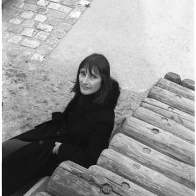 Black-and-white photograph of Eva Figes, looking up at the camera and sitting beside log stairs, 14 October
            1980.