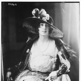 Black and white, three-quarter-length photo of Rosita Forbes by Mendoza. She is seated, gazing at the viewer, in a dark hat with wide brim and large artificial flowers, and a fur wrap thrown over an open jacket and light dress with a full, gauzy or lacy skirt. Library of Congress.