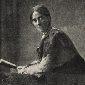 Black and white photo of Isabella Ford in 1924, sitting reading a book with a telephone beside her. Her body is in profile but she turns her head to face the camera. Her dark hair is parted in the middle and pulled back.