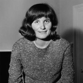 Black-and-white portrait photo of Margaret Forster, looking at the camera and leaning on what could be the back of a couch, 30 January 1964. 