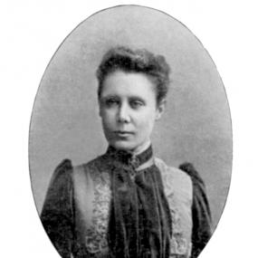 Black and white photo of Jessie Fothergill. Her hair is short and brushed back. She is wearing a dress with frilly collar, a waistcoat over it, and a brooch on her collar. Her expression is stern.