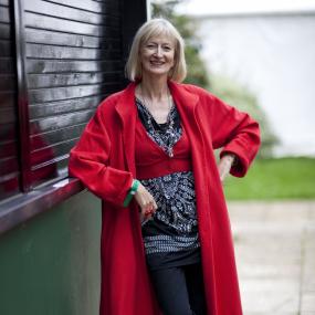 Head-to-toe colour photo portrait of Maggie Gee at the Cheltenham Literature Festival, 9 October 2010. She is wearing a black and red outfit,
            complete with red-laced shoes, a red coat, and red rings on her fingers. The photo is taken outside. 