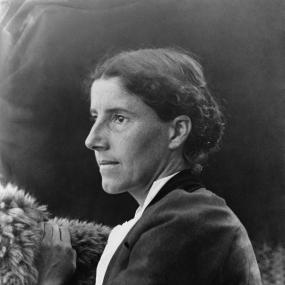 Black and white photograph of Charlotte Perkins Gilman, shown in profile, from the torso up, with one hand resting on a fur wrap. She wears a dark jacket over a white shirt and her wavy hair is done in a loose bun.