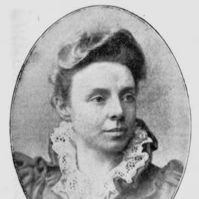 Black and white, oval-framed photo of Katharine Bruce Glasier from the neck up. Her hair is up, with a wave in front going towards the left. The dress she is wearing has a large standing collar lined with lace. Below is written, the first line in capitals: "Katharine St. John Conway / Mrs Bruce Glasier".