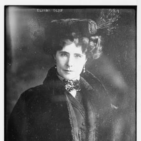 Black and white photo of Elinor Glyn from the waist up. She is wearing drop ear-rings, a fur hat, and a fur-trimmed coat over a suit. Her hands are in a fur muff. Her hair is short, in luxuriant curls. Her gaze is directed at the viewer.