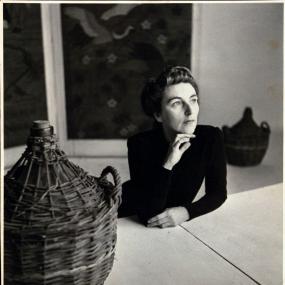 Black-and-white photo of Rumer Godden. She is posing at a table with her hand on her chin. There are two wicker baskets in the image, one in
            the foreground and one in the back. 