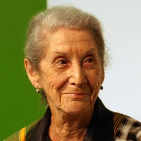Colour photo of Nadine Gordimer. Her grey hair is brushed back; her lined face has an impish smile. She is wearing an open-necked shirt which is checked in various colours and styles, with stud earrings and a Swedish flag lapel pin.