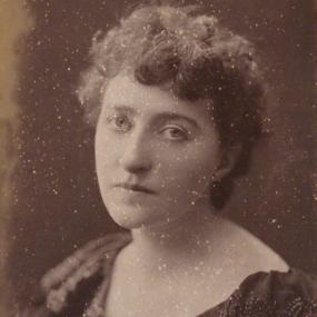 Sepia albumen print (flecked with white) of Sarah Grand by Hayman Seleg Mendelssohn, 1894. Grand is seen from the shoulders up, at a slight turn, wearing a dark dress with beaded detail at the neck. She has short, dark, curly hair.