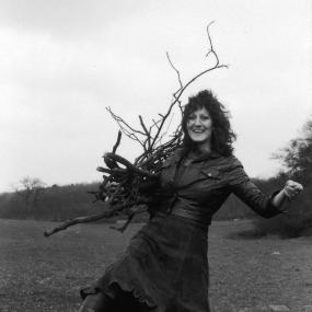 Black-and-white photo of Germaine Greer, looking at the camera and holding a tall bunch of sticks as she stretches one leg over a fence, March
            1971.