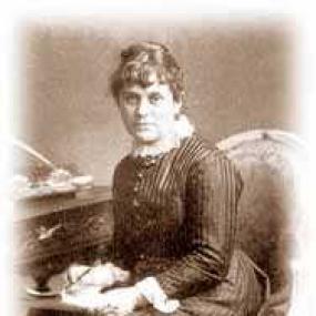Low resolution sepia photo of Kate Greenaway. She is wearing a striped, buttoned dress with a white collar, sitting in a chair with pen and paper in her hands, beside a desk with, apparently, more writing materials. Her hair is in short curls in front with a bun behind.