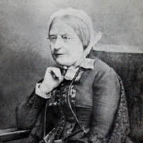 Low-resolution black-and-white photo of Maria Grey sitting in a chair,holding a book. She has a white cap tied round her head and she is wearing glasses.  Her dress is buttoned up the front and topped by a short jacket.