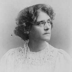 Black and white photograph of Beatrice Harraden, shown from the shoulders up. She has her head turned and appears to be looking off intently into the distance. She wears a light dress with flower detailing around the neck, and she has short curly hair and small wire-rimmed spectacles perched on the end of her nose.