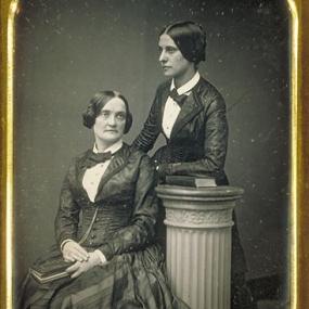 Black and white photograph of Matilda Hays (standing on the right) with Charlotte Cushman (sitting on the left). Matilda Hays is wearing a fancy vest and skirt, with a bowtie around her neck. Her hair is parted in the middle and tied back. Her left arm is resting on a book on top of a doric pillar with floral design on the sides near the top. Charlotte Cushman is wearing a very similar outfit except her skirt has some striped design on it. Her hair is parted in the middle, but it is tied up in a double bun.