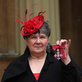 Colour photo of Susan Hill posing with her Commander of the British Empire (CBE) medal at Buckingham Palace, after her investiture ceremony on
            13 December 2012. She wears a bright red fascinator hat with feathers and a red rose. 