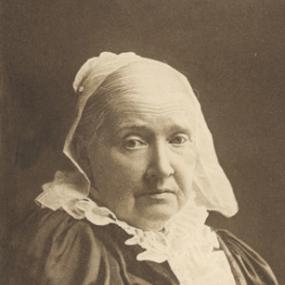 Black and white half-length photograph of Julia Ward Howe, seated and turning a little towards the camera. She wears an ample  dark dress with white ruffles at the throat and front, and a white cap over her ears and her grey hair.